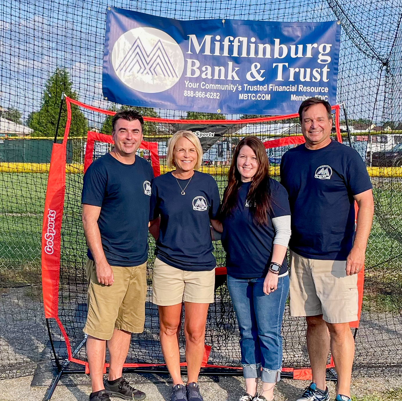 A group of 4 employees from Mifflinburg Bank stand outside in front of a baseball cage in the summertime at a community event.